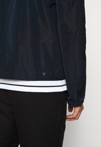 Navy blue, lightweight jacket with a smooth texture. Features a snap closure at the hem and a wrist cuff with a button. Striped shirt underneath.