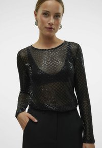Schwarzes Mesh-Top mit einem Rautenmuster aus Pailletten, langen Ärmeln und einem runden Ausschnitt, kombiniert mit hochtaillierten schwarzen Hosen.