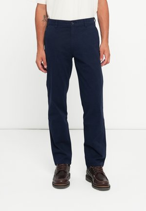 Navy blue trousers made of cotton with a straight cut, featuring a front button closure and side pockets. Worn with brown lace-up shoes.
