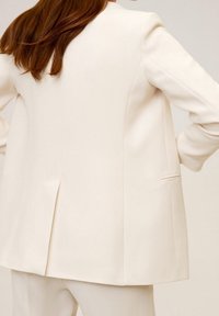 Blazer blanc sur mesure avec une fente unique à l'arrière, des épaules structurées et une texture élégante. Détail subtil de poche latérale.