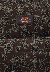 Tissu marron avec motif floral et feuillagé noir et beige, présentant un patch rectangulaire avec le logo Dickies cousu au centre.
