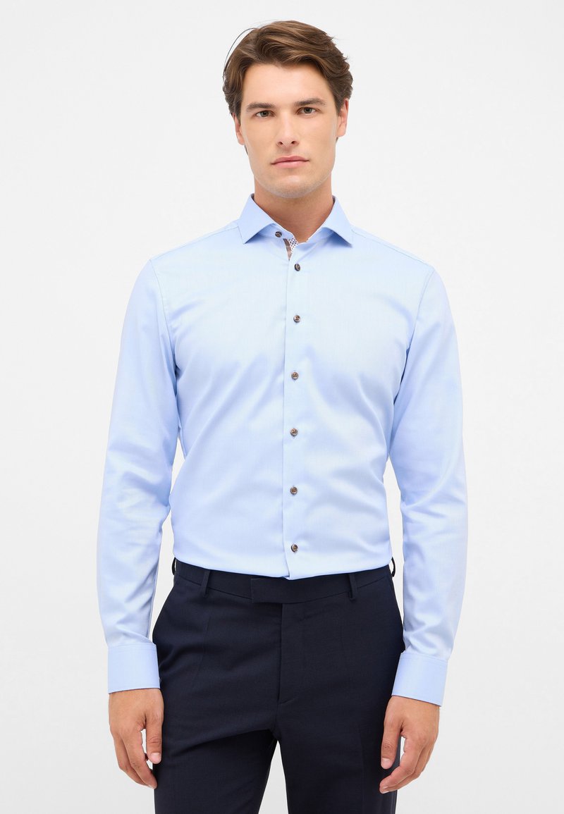 Camicia azzurro chiaro con maniche lunghe, caratterizzata da un sottile motivo a righe e bottoni marroni, abbinata a pantaloni scuri.