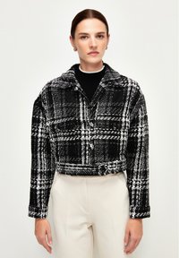 Chaqueta de cuadros recortada en blanco y negro, hecha de tejido texturizado con un cuello, dos bolsillos frontales y cierres de botón.