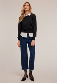 Un pull noir sur une chemise col blanc, associé à un jean bleu à larges jambes avec des ourlets effilochés et des chaussettes en résille noires avec des talons.