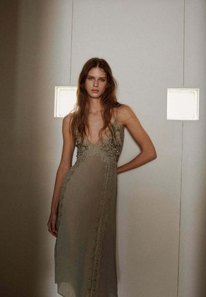 Jeune femme aux longs cheveux portant une robe beige sans manches avec des détails en dentelle et perles, debout contre un mur blanc avec deux fenêtres carrées.