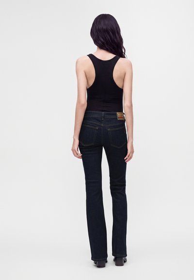 Femme aux longs cheveux foncés portant un haut sans manches noir et un jean bleu foncé, debout, de dos, sur un fond blanc uni.