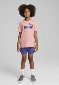Camiseta de algodón rosa con logotipo azul de "PUMA", combinada con pantalones cortos azul marino y zapatillas grises. Calcetines hasta el tobillo visibles. Postura recta contra un fondo gris.