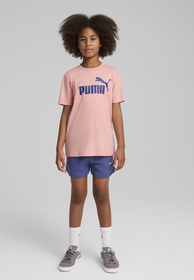 Camiseta de algodón rosa con logotipo azul de "PUMA", combinada con pantalones cortos azul marino y zapatillas grises. Calcetines hasta el tobillo visibles. Postura recta contra un fondo gris.