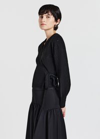 Maglione avvolgente nero lavorato a maglia con un motivo testurizzato, maniche lunghe e un fiocco in vita, abbinato a una gonna nera.