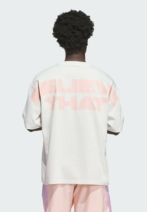 Person med krøllet hår set bagfra iført en hvid oversized T-shirt med store lyserøde bogstaver, der delvist lyder "BELIEVE THAT".