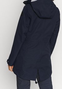 Chaqueta impermeable azul marino con capucha, cintura ajustable con cordón y costuras visibles. Presenta un logo en la manga izquierda y un diseño de espalda partida.