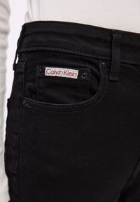 Černé džíny z denimu s úzkým střihem, vybavené pěti kapsami, viditelným šitím a červenobílou etiketou Calvin Klein na zadní kapse.