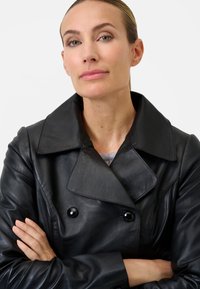 Black leather trench coat with large collar, double-breasted buttons, and smooth texture. Worn over a grey top, arms crossed.
