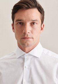 Biała koszula z kołnierzykiem o gładkiej fakturze, z zapięciem na guziki i klasycznym krojem. Gładki materiał, bez wzorów i akcentów.