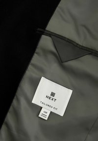 Fekete öltönydzseki belseje, sima szürke béléssel, háromszögletű zsebbel és egy fehér márkacímkével, amelyen a "NEXT TAILORED FIT 40R" olvasható.