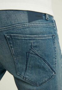 Modré džíny z denimu s upnutým střihem, které mají na zadních kapsách tmavě modrou koženou nášivku s nápisem "CHASIN'" a bílým prošíváním.