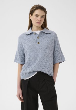 Naine, kes kannab helesinist lühikeste varrukatega kootud polosärki nööpidega, kokku sobitatud mustade kõrge vöökohaga volditud pükstega, seisab valge tausta ees.