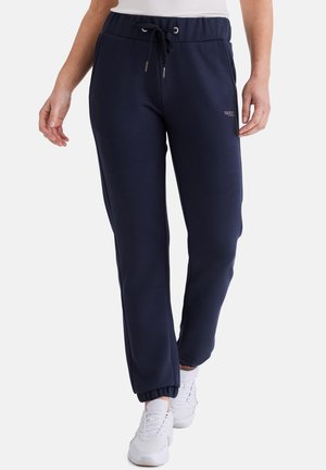 Frau trägt marineblaue Jogginghosen mit elastischen Bündchen und Kordelzug in der Taille, kombiniert mit weißen Sneakern und einem weißen Oberteil.