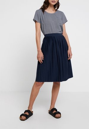 Woman wearing a short-sleeve patterned top tucked into a navy pleated knee-length skirt with black buckle sandals, standing with one leg forward.