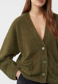 Kardigan w oliwkowym odcieniu zieleni, puszysty, z głębokim dekoltem w kształcie litery V, wyposażony w duże guziki i boczne kieszenie. Miękka faktura z luźnymi rękawami.