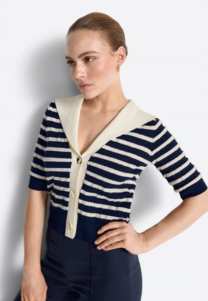 Naine, kellel on juuksed seljataga seotud, seljas tumesinine ja valge triibuline lühikese varrukaga kardigan suure kraega ja kuldsete nuppudega, poseerib käega puusal.