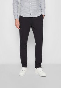 Herren Slim-Fit schwarze Hosen aus leichtem Stoff, mit Seitentaschen und glatter Textur. Getragen mit einem hell gestreiften Hemd und weißen Sneakern.