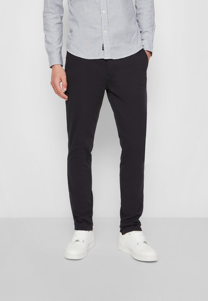 Herren Slim-Fit schwarze Hosen aus leichtem Stoff, mit Seitentaschen und glatter Textur. Getragen mit einem hell gestreiften Hemd und weißen Sneakern.
