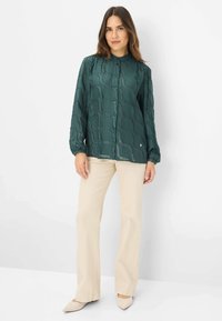 Green blouse made of textured fabric with a wave pattern, featuring a button-up front and long sleeves, paired with beige pants and pointed shoes.