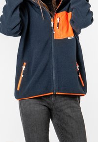 Fleecepusero tummansinisessä värissä oranssilla rintataskulla. Varustettu vetoketjullisilla taskuilla ja kaksisuuntaisella vetoketjulla. Pehmeä pinta, rento malli.