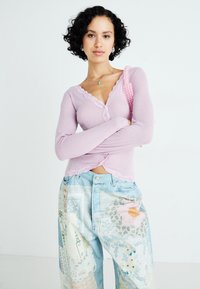 Lichtroze gerekte top met lange mouwen, afgewerkt met kant en knopen; gecombineerd met patchwork spijkerbroek in het blauw met bloemen- en kantpatronen.