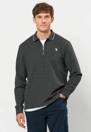 Hombre con polo gris oscuro de manga larga con cremallera y pequeño logo blanco, combinado con pantalones azul oscuro, de pie con una mano en el bolsillo.