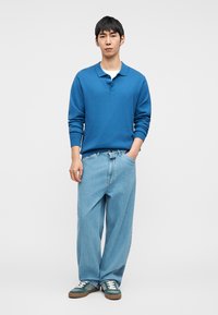 Νεαρός άνδρας που στέκεται, φοράει μπλε πόλο με μακριά μανίκια, ανοιχτόχρωμο τζιν με φαρδιά μπατζάκια, πράσινα αθλητικά παπούτσια, με το ένα χέρι στην τσέπη.