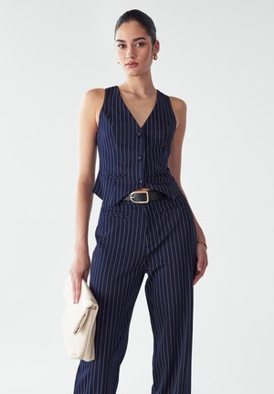 Marineblaues Nadelstreifen-Outfit bestehend aus einem ärmellosen Oberteil mit Peplum-Design und passenden Hosen, kombiniert mit einer elfenbeinfarbenen Clutch.