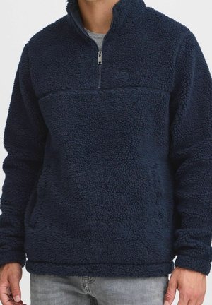 Mężczyzna ubrany w granatowy, teksturowany polar z zamkiem do ćwiartki oraz szarą koszulę pod spodem, ręce swobodnie opadające wzdłuż ciała.