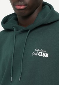 Zelená mikina s kapucňou vyrobená z jemného materiálu, s bielym nápisom: "Chalet Royale Ski CLUB" na prednej strane.
