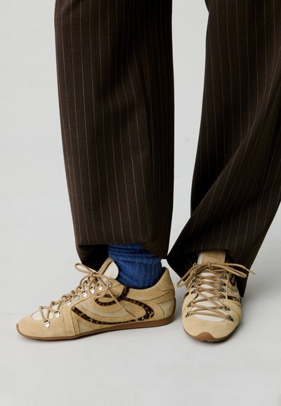 Sneaker in suede beige con dettagli in stampa animale marrone, punta tonda e design con lacci. Indossati con pantaloni scuri a righe e calzini blu.
