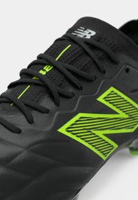 Melna New Balance futbola zābaku ar neona zaļu logotipu un melnām auklām uz baltas fona.