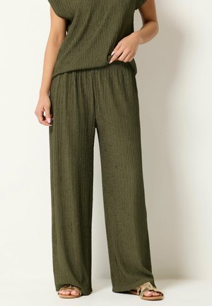 Mujer con pantalones de punto acanalados de pierna ancha en verde oliva combinados con un top a juego y sandalias doradas de punta abierta, de pie frente a un fondo liso.