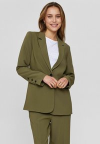 Oliivroheline bleiser, millel on kohandatud lõige, ühe nupuga kinnitamine, sakilised kraed ja nelja nupuga varrukad, kantud valge tekstuuriga topi üle.