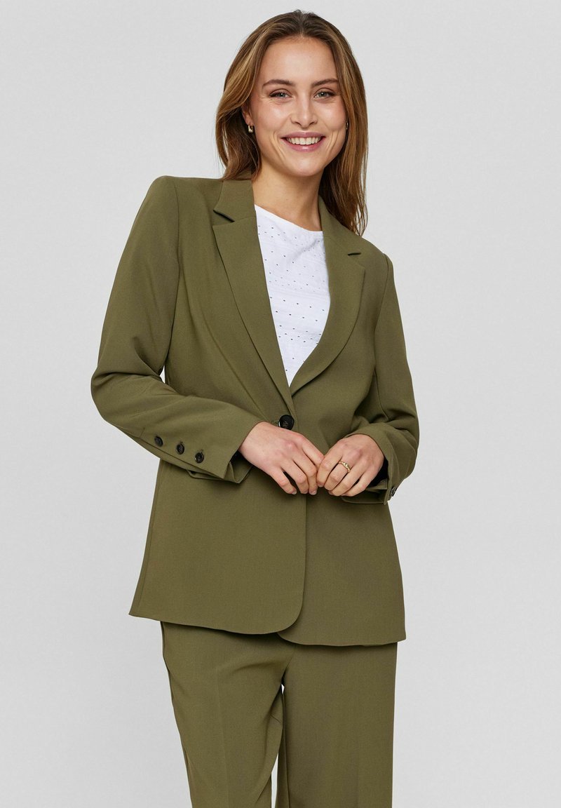 Oliivroheline bleiser, millel on kohandatud lõige, ühe nupuga kinnitamine, sakilised kraed ja nelja nupuga varrukad, kantud valge tekstuuriga topi üle.