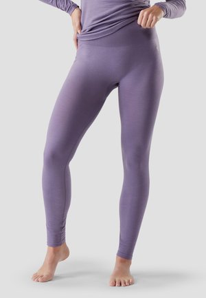 Osoba ubrana w dopasowane lawendowe legginsy i pasującą bluzkę z długim rękawem, stojąca boso na jednolitym jasnym tle.