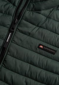 Mørkegrøn quiltet jakke med sort lynlås med "SUPERDRY®"-logo på lynlåstrækker og brystpatch.