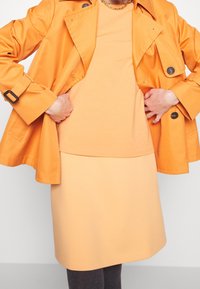 Un manteau trench orange sur un top et une jupe pêche, en tissu lisse et au design simple, avec un col rond et des coutures subtiles.