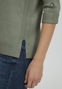 Un pull en maille vert clair avec des manches au niveau des coudes, présentant une fente à l'ourlet et un accent en bouton sur le poignet roulé. Porté par-dessus un jean noir.