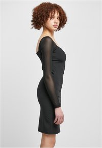 Robe noire ajustée avec de longues manches transparentes, encolure ronde et texture lisse. Conception longueur genoux avec une silhouette cintrée.