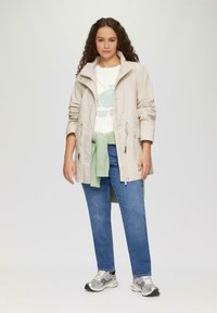 Jasno beżowa kurtka z zapięciem na zamek, dwoma kieszeniami z przodu i regulowanymi rękawami; w zestawie z białym t-shirtem z grafiką i niebieskimi dżinsami.