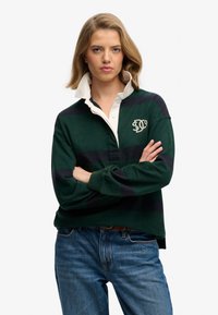 Femme aux bras croisés portant un polo rayé vert et bleu marine avec un col blanc et un jean bleu, debout devant un fond blanc.