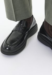 Mocasines de cuero negro con un acabado liso y una correa decorativa, que cuentan con una gruesa suela de goma negra. Combinados con calcetines marrones de canalé y pantalones grises.