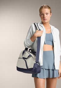 Harmaa ja laivastonsininen duffel-laukku, jossa on jäsennelty muotoilu, yläkahvat, säädettävä olkahihna ja etutaskussa vetoketju.