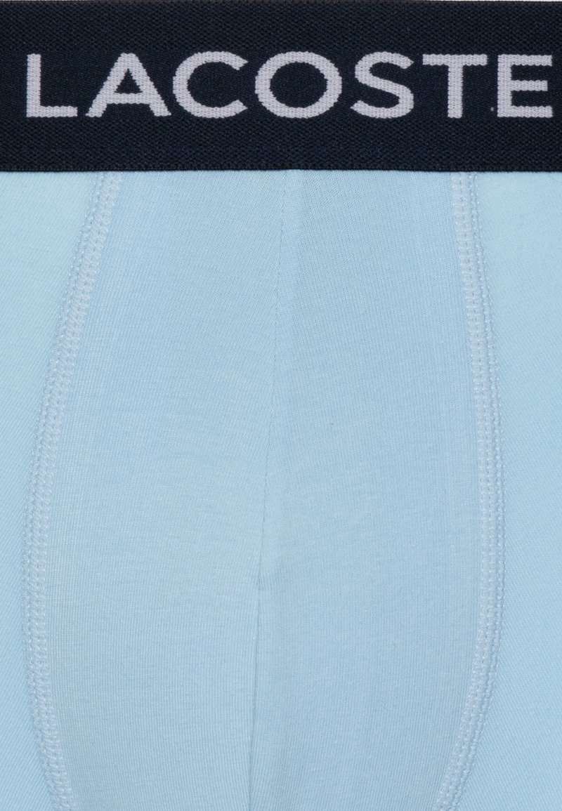 Lichtblauwe katoenen boxershorts met een marineblauwe elastische tailleband met het "LACOSTE" logo in grijs. Naaddetailing is zichtbaar.